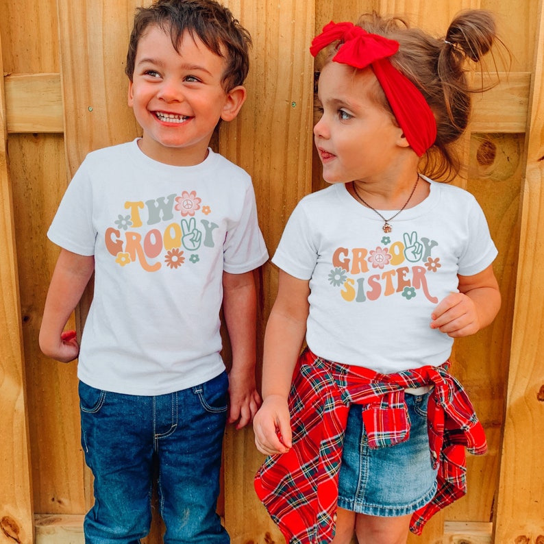 May include: Two children wearing white t-shirts with a groovy theme. The boy's shirt says "Two Groovy" and the girl's shirt says "Groovy Sister". Both shirts feature peace signs and flowers.