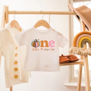 Puede incluir: Camiseta blanca de manga corta con el texto "one" en naranja, rosa y azul con un gr&aacute;fico de calabaza y el texto "Little Pumpkin" debajo.