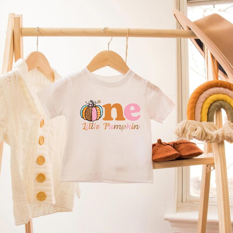 Puede incluir: Camiseta blanca de manga corta con el texto "one" en naranja, rosa y azul con un gr&aacute;fico de calabaza y el texto "Little Pumpkin" debajo.
