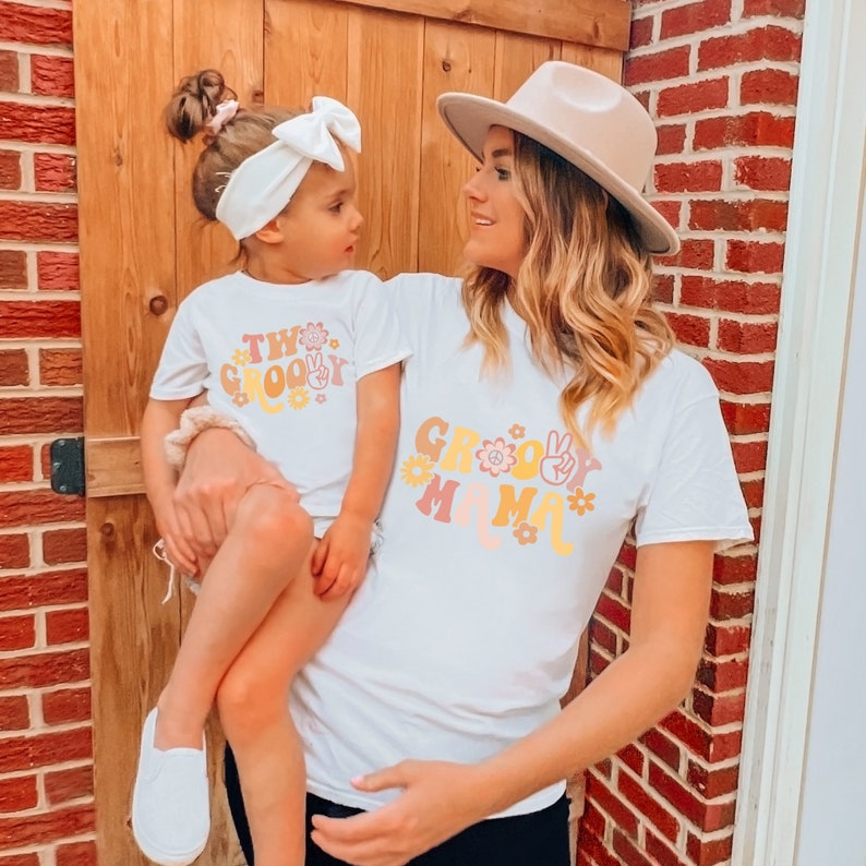 May include: A white t-shirt with the text "Groovy Mama" in a retro font with flowers and peace signs. A child is wearing a white t-shirt with the text "Two Groovy" in a retro font with flowers and peace signs.