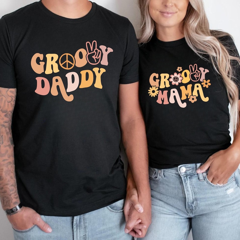 May include: Two black t-shirts with a retro groovy font design. The shirt on the left says "Groovy Daddy" with a peace sign and a peace sign hand gesture. The shirt on the right says "Groovy Mama" with flowers and a peace sign hand gesture.