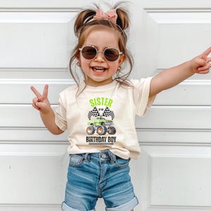 Puede incluir: Una ni&ntilde;a lleva una camiseta color crema con la frase "SISTER OF THE BIRTHDAY BOY" y un dise&ntilde;o de cami&oacute;n monstruo verde. La ni&ntilde;a lleva pantalones cortos vaqueros y gafas de sol. La imagen muestra a una ni&ntilde;a feliz haciendo el signo de la paz.
