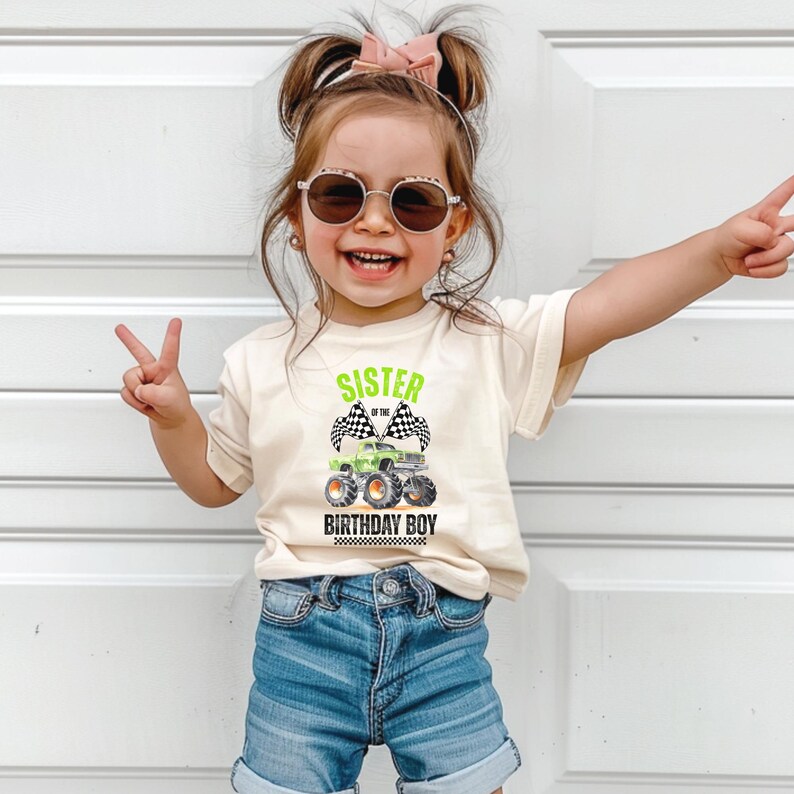 Puede incluir: Una ni&ntilde;a lleva una camiseta color crema con la frase "SISTER OF THE BIRTHDAY BOY" y un dise&ntilde;o de cami&oacute;n monstruo verde. La ni&ntilde;a lleva pantalones cortos vaqueros y gafas de sol. La imagen muestra a una ni&ntilde;a feliz haciendo el signo de la paz.