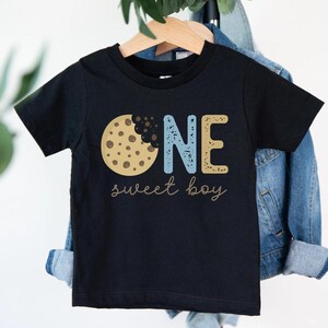 May include: Black toddler t-shirt with a cookie graphic and the words "ONE sweet boy." The word "ONE" is in blue and gold block letters. The cookie is brown with chocolate chips.