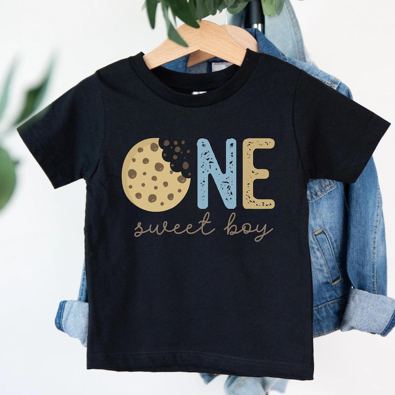 May include: Black toddler t-shirt with a cookie graphic and the words "ONE sweet boy." The word "ONE" is in blue and gold block letters. The cookie is brown with chocolate chips.