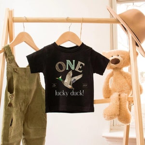 May include: Black short-sleeved t-shirt with a green, brown and white "ONE" graphic and a flying duck illustration. The text below the graphic reads "Est. 2024 lucky duck!"