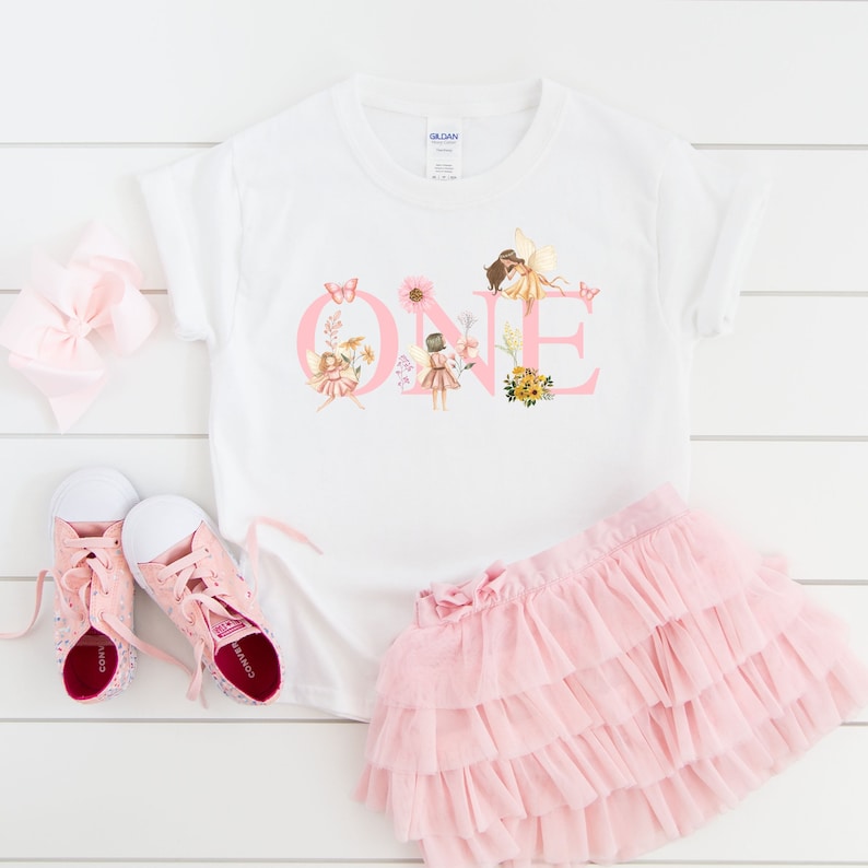 May include: A white short-sleeved t-shirt featuring the pink text ONE decorated with illustrations of fairies, flowers, and butterflies. The set includes a light pink tiered tulle skirt, a pair of pink canvas sneakers, and a pink hair bow.