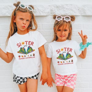 Może przedstawiać: Dwie młode dziewczyny w pasujących białych koszulkach z napisem "Sister of the Hungry Caterpillar" i kolorowym motywem motyla. Jedna dziewczyna nosi czarno-białe szorty w kratę, druga r&oacute;żowo-białe szorty w kratę.