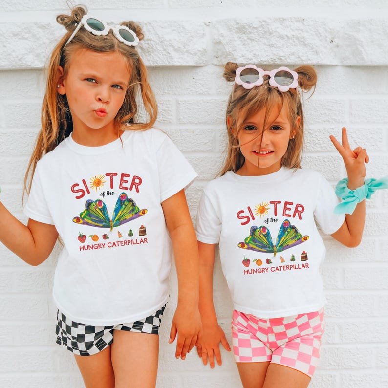 Może przedstawiać: Dwie młode dziewczyny w pasujących białych koszulkach z napisem "Sister of the Hungry Caterpillar" i kolorowym motywem motyla. Jedna dziewczyna nosi czarno-białe szorty w kratę, druga r&oacute;żowo-białe szorty w kratę.