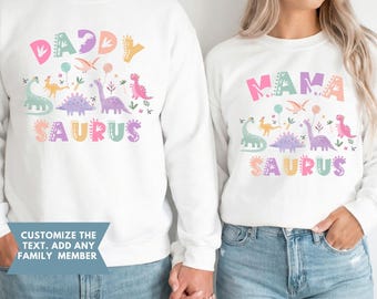 May include: White sweatshirts with colorful dinosaur graphics and the words "DADDY SAURUS" and "MAMA SAURUS". The sweatshirts have long sleeves and are worn with blue jeans. The text "CUSTOMIZE THE TEXT. ADD ANY FAMILY MEMBER" is also visible.