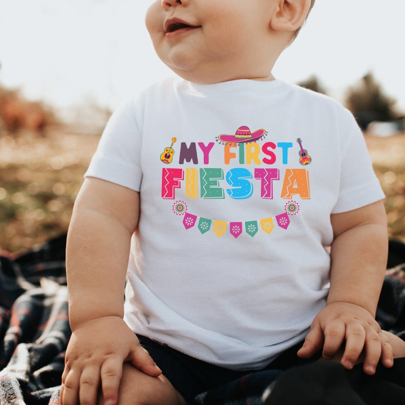 K&ouml;nnte beinhalten: Ein wei&szlig;es T-Shirt mit dem Text "My First Fiesta" in bunten Buchstaben. Der Text ist mit einem Sombrero, einer Gitarre und bunten Flaggen verziert.