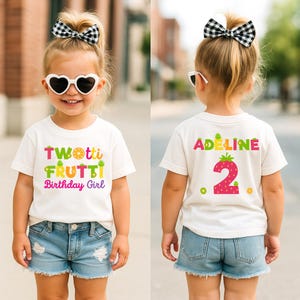 May include: A young child wearing a white t-shirt with the text "Twotti Frutti Birthday Girl" in colorful letters. The child is also wearing denim shorts, heart-shaped sunglasses, and a black and white checkered bow.