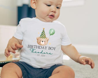 May include: White t-shirt with a watercolor illustration of a teddy bear wearing a party hat and holding a green balloon. The text reads "BIRTHDAY BOY Theodore - EST. 2024". The shirt is worn by a baby.