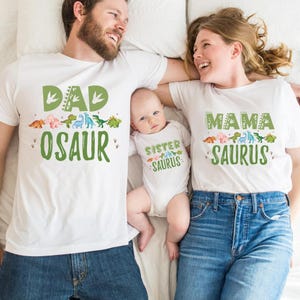 May include: A family matching set of white t-shirts with dinosaur graphics and text. The dad's shirt says "Dad-osaur", the mom's shirt says "Mama-saurus", and the baby's shirt says "Sister-saurus".