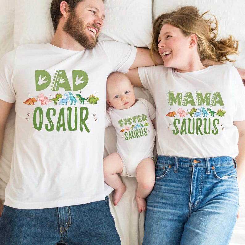 May include: A family matching set of white t-shirts with dinosaur graphics and text. The dad's shirt says "Dad-osaur", the mom's shirt says "Mama-saurus", and the baby's shirt says "Sister-saurus".