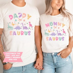 May include: Matching white t-shirts with dinosaur graphics and the words "Daddy Saurus" and "Mommy Saurus" in pastel colours. The shirts also feature the text "Customize the text. Add any family member and age."