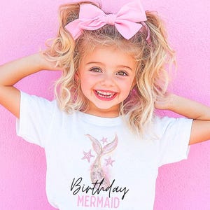 May include: A young girl with blonde hair and a pink bow smiles, wearing a white t-shirt with a mermaid tail graphic and the words "Birthday Mermaid". The background is a solid pink color.