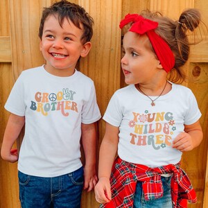 May include: Two children wearing white t-shirts with colorful retro-style text. The boy's shirt says "Groovy Brother" and the girl's shirt says "Young Wild & Three".