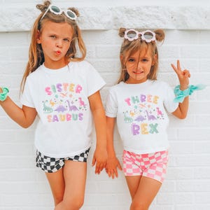 May include: Two young girls in white t-shirts with dinosaur designs and text. One shirt reads "Sister Saurus" and the other "Three Rex." They are also wearing shorts, one with a black and white checkerboard pattern and the other with a pink and white checkerboard pattern.