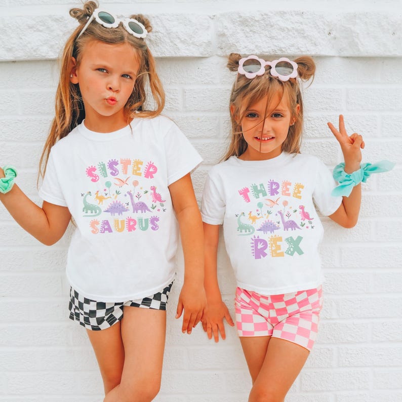 May include: Two young girls in white t-shirts with dinosaur designs and text. One shirt reads "Sister Saurus" and the other "Three Rex." They are also wearing shorts, one with a black and white checkerboard pattern and the other with a pink and white checkerboard pattern.