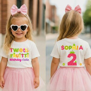 May include: A white t-shirt featuring the text TWOTTI FRUITTI Birthday Cutie on the front and the name SOPHIA with a large number 2 on the back, paired with a light pink tulle skirt, a matching pink hair bow, and white heart-shaped sunglasses.