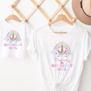 K&ouml;nnte beinhalten: Zwei wei&szlig;e T-Shirts mit einem Regenbogen- und Einhorn-Design. Das kleinere Shirt sagt "Birthday Girl". Das gr&ouml;&szlig;ere Shirt sagt "Mommy of the Birthday Girl".