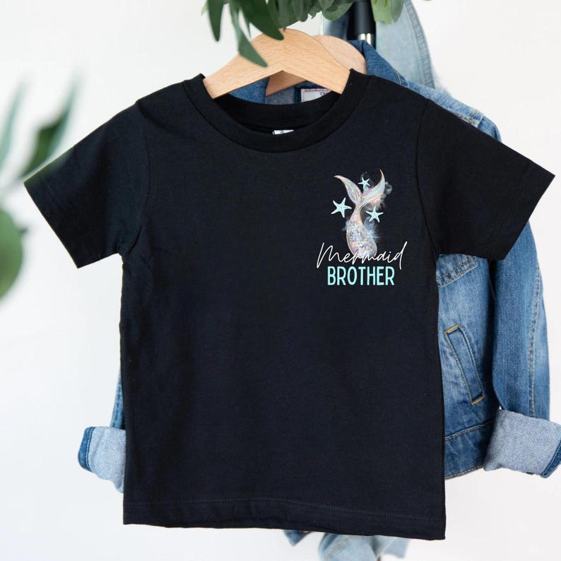 May include: Black short-sleeved t-shirt with a mermaid tail and star graphic. The text "Mermaid Brother" is printed below the graphic in a script font. The shirt is hanging on a wooden hanger.