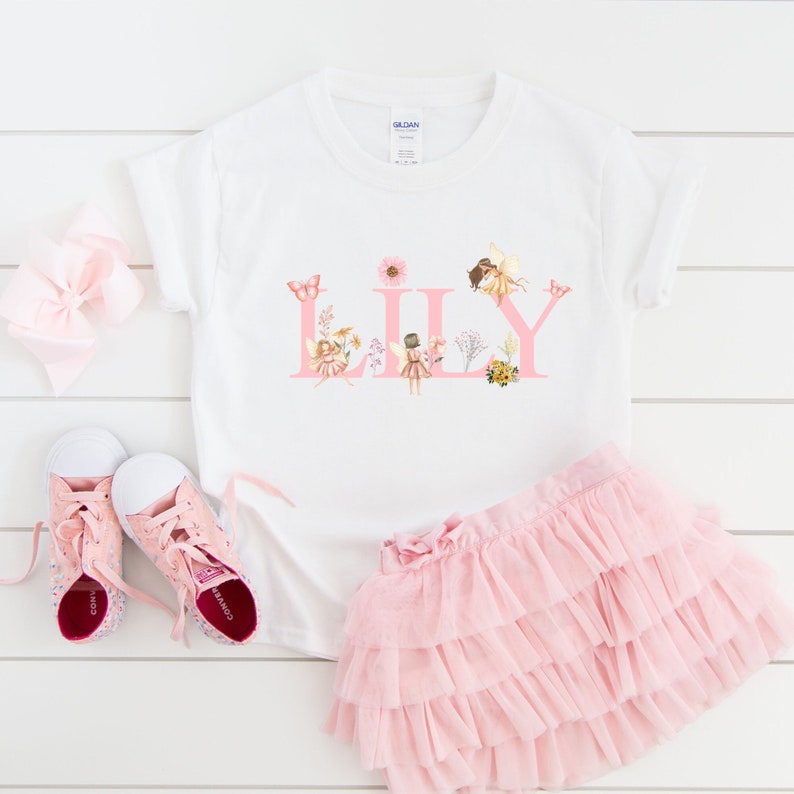 May include: A white short-sleeve t-shirt featuring the name LILY in large pink letters decorated with fairy illustrations, flowers, and butterflies. The set includes a light pink tiered tulle skirt and a pair of pink canvas sneakers.