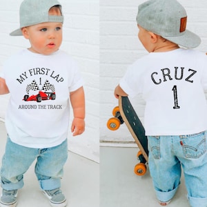 Peut inclure: T-shirt blanc avec l'inscription "MY FIRST LAP" et un dessin de voiture de course. Le dos du t-shirt porte le nom "CRUZ" et le numéro "1". L'enfant porte un jean bleu et une casquette de baseball grise.