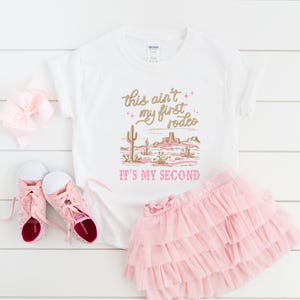 May include: White t-shirt with pink and gold text that reads "This ain't my first rodeo. It's my second."  The shirt features a desert scene with cacti and mountains.