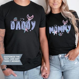 May include: Two black t-shirts with a mermaid tail design and the words "Daddy" and "Mommy" in purple. The shirts are being worn by two people holding hands.