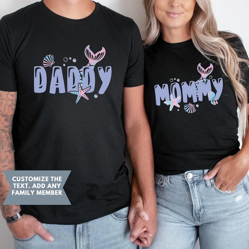 May include: Two black t-shirts with a mermaid tail design and the words "Daddy" and "Mommy" in purple. The shirts are being worn by two people holding hands.