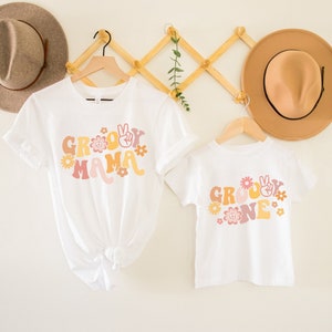 May include: Two white t-shirts with a groovy theme. The larger shirt says "Groovy Mama" in a retro font with peace signs and flowers. The smaller shirt says "Groovy One" in the same font with peace signs and flowers.