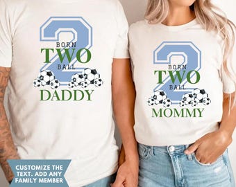 May include: Two white t-shirts featuring a large light blue number two, the text BORN TWO BALL, and soccer ball graphics. One shirt displays the word DADDY in green, while the other displays the word MOMMY in green below the soccer ball design.