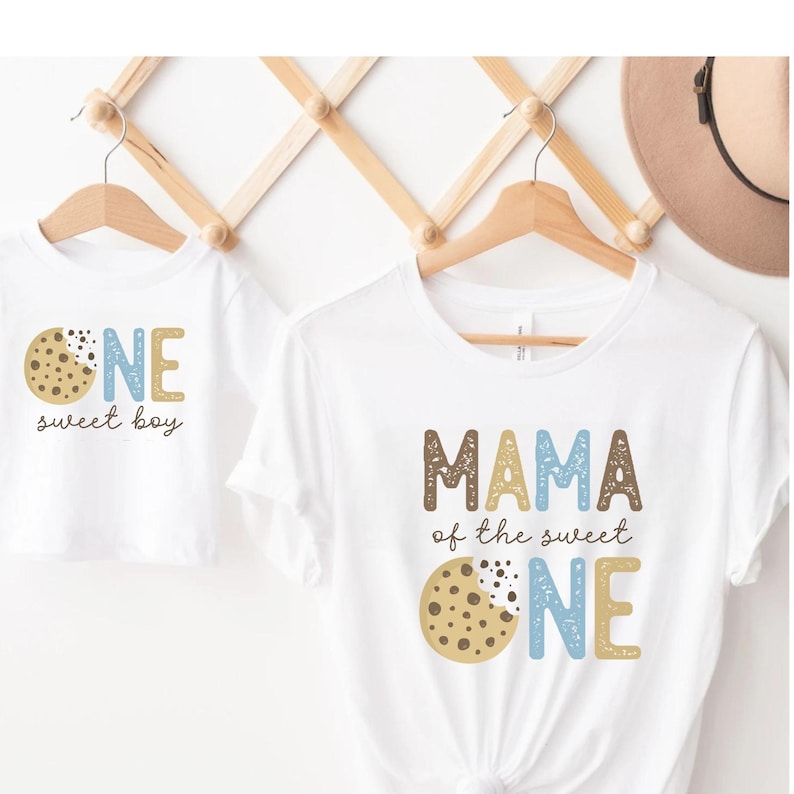 May include: Two white t-shirts with matching designs. One says "ONE sweet boy" with a cookie graphic. The other says "MAMA of the sweet ONE" with a cookie graphic. Both shirts are hanging on wooden hangers.