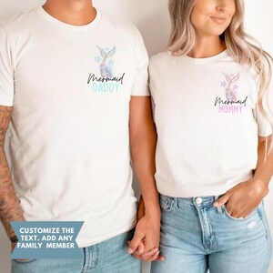 May include: Two off-white t-shirts with mermaid tail and star graphics. One shirt says "Mermaid Daddy" in blue, the other says "Mermaid Mommy" in pink. The shirts are worn by two people. The text "Customize the text. Add any family member" is at the bottom.