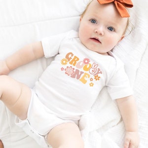 May include: A white baby onesie with the text "Groovy One" in orange, yellow, and pink letters. The text is decorated with peace signs and flowers.