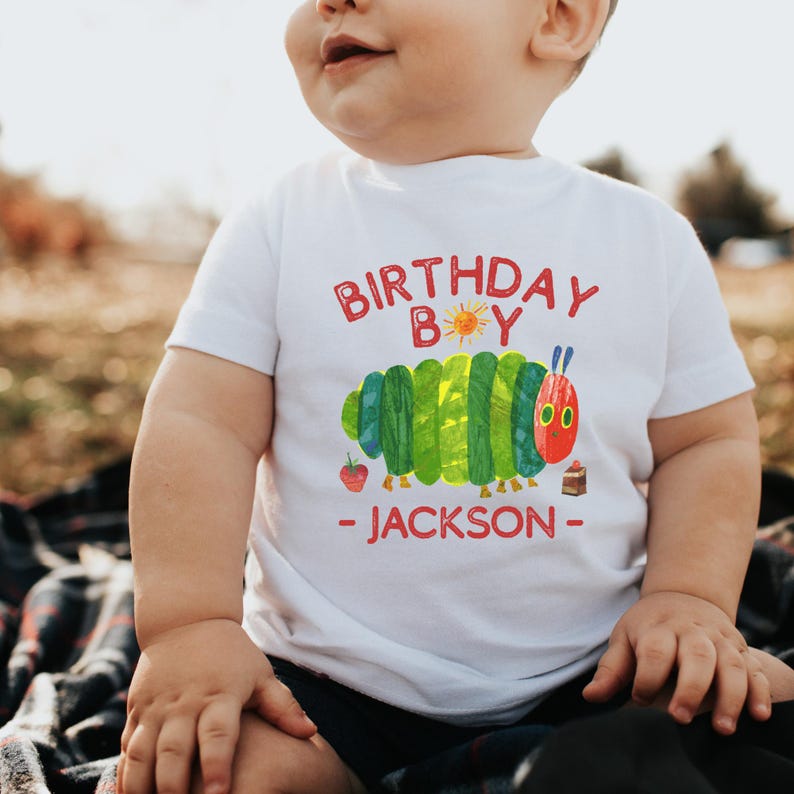 Może przedstawiać: Biała koszulka z kolorową grafiką gąsienicy i napisem "BIRTHDAY BOY - JACKSON" w kolorze czerwonym. Gąsienica jest zielono-ż&oacute;łta z czerwoną głową. Występują r&oacute;wnież truskawka i ciasto.