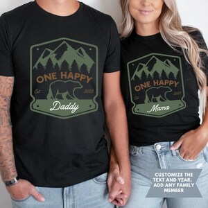 May include: Two black t-shirts with a green mountain and bear design. The text on the shirts reads "One Happy" with the year "2023" and the words "Daddy" and "Mama".