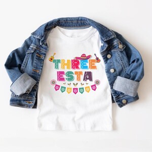 Peut inclure: T-shirt blanc avec un graphisme color&eacute; "THREE ESTA". Le graphisme repr&eacute;sente un sombrero, des maracas et des guitares. Le t-shirt est d&eacute;cor&eacute; d'une guirlande de drapeaux color&eacute;s.