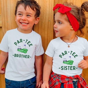 May include: Two children wearing white t-shirts with a golf-themed design. The shirts feature the text "PAR-TEE" above a golf cart, golf ball, and flag, with "BROTHER" and "SISTER" below. The background is a wooden fence.