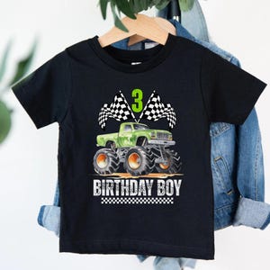 Puede incluir: Camiseta negra con un dise&ntilde;o de cami&oacute;n monstruo verde, ruedas naranjas, banderas a cuadros y el n&uacute;mero 3. El texto "BIRTHDAY BOY" est&aacute; impreso debajo. Una divertida camiseta gr&aacute;fica para una celebraci&oacute;n de tercer cumplea&ntilde;os.