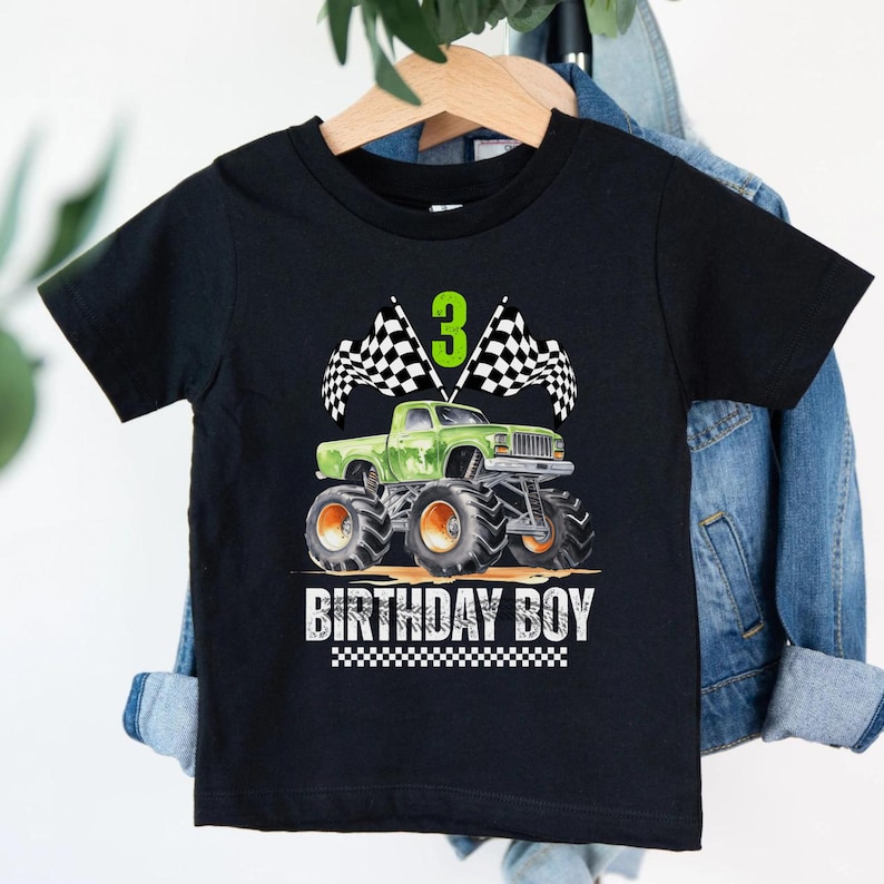 Puede incluir: Camiseta negra con un dise&ntilde;o de cami&oacute;n monstruo verde, ruedas naranjas, banderas a cuadros y el n&uacute;mero 3. El texto "BIRTHDAY BOY" est&aacute; impreso debajo. Una divertida camiseta gr&aacute;fica para una celebraci&oacute;n de tercer cumplea&ntilde;os.