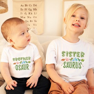 May include: Two children wearing white t-shirts with dinosaur graphics. The boy's shirt says "Brother Osaur" and the girl's shirt says "Sister Saurus".