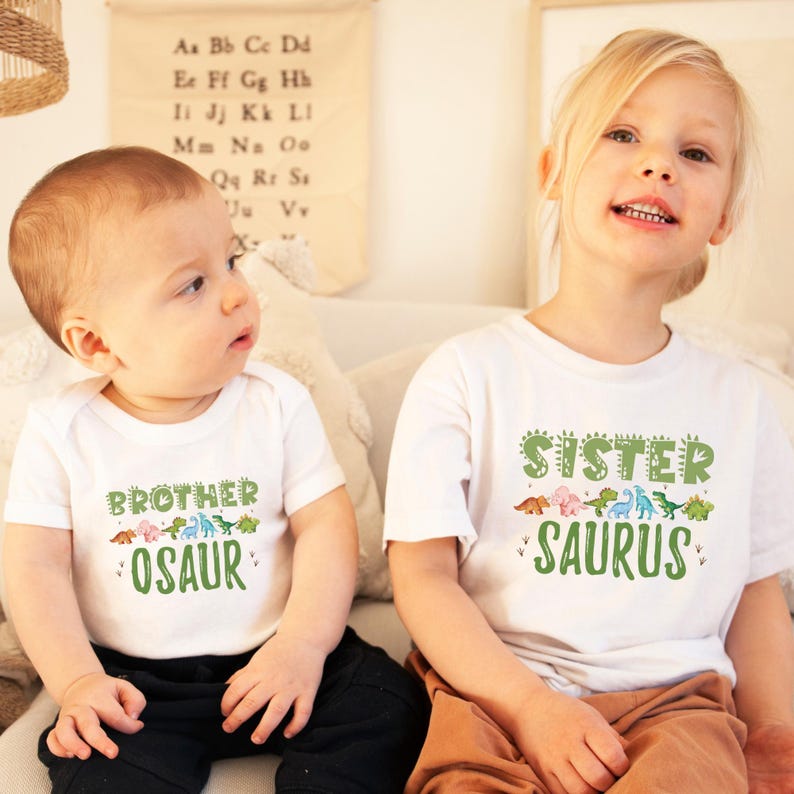 May include: Two children wearing white t-shirts with dinosaur graphics. The boy's shirt says "Brother Osaur" and the girl's shirt says "Sister Saurus".
