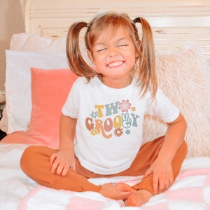 May include: A young child wearing a white t-shirt with the text "Two Groovy" in a retro font with peace signs and flowers. The child is sitting on a bed with a pink and white blanket.