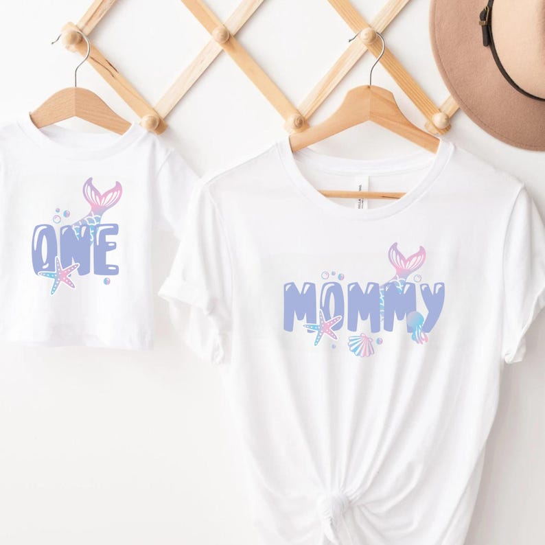 May include: Two white t-shirts with a mermaid tail design. The smaller shirt says "ONE" in blue with a mermaid tail and a starfish. The larger shirt says "MOMMY" in blue with a mermaid tail, a starfish, and seashells.