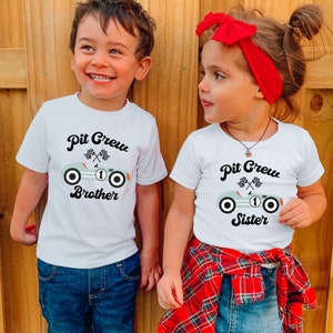 May include: Two children wearing white t-shirts with a blue and white race car graphic and the text "Pit Crew" on them. The boy's shirt says "Brother" and the girl's shirt says "Sister".