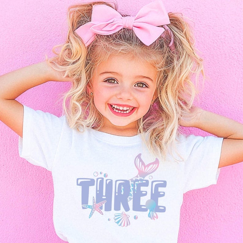 May include: A young child wearing a white t-shirt with the word "THREE" in blue, accompanied by a mermaid tail and sea-themed graphics. The child has blonde hair styled with a pink bow, smiling in front of a pink background.