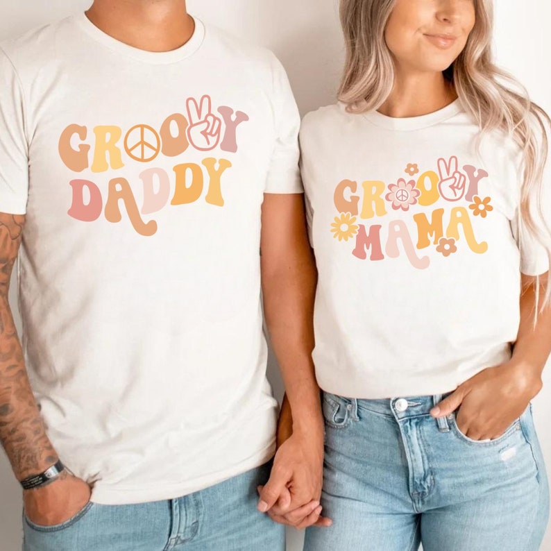 May include: Two white t-shirts with a retro groovy font design. The shirt on the left says "Groovy Daddy" and the shirt on the right says "Groovy Mama". Both shirts feature a peace sign and flowers.